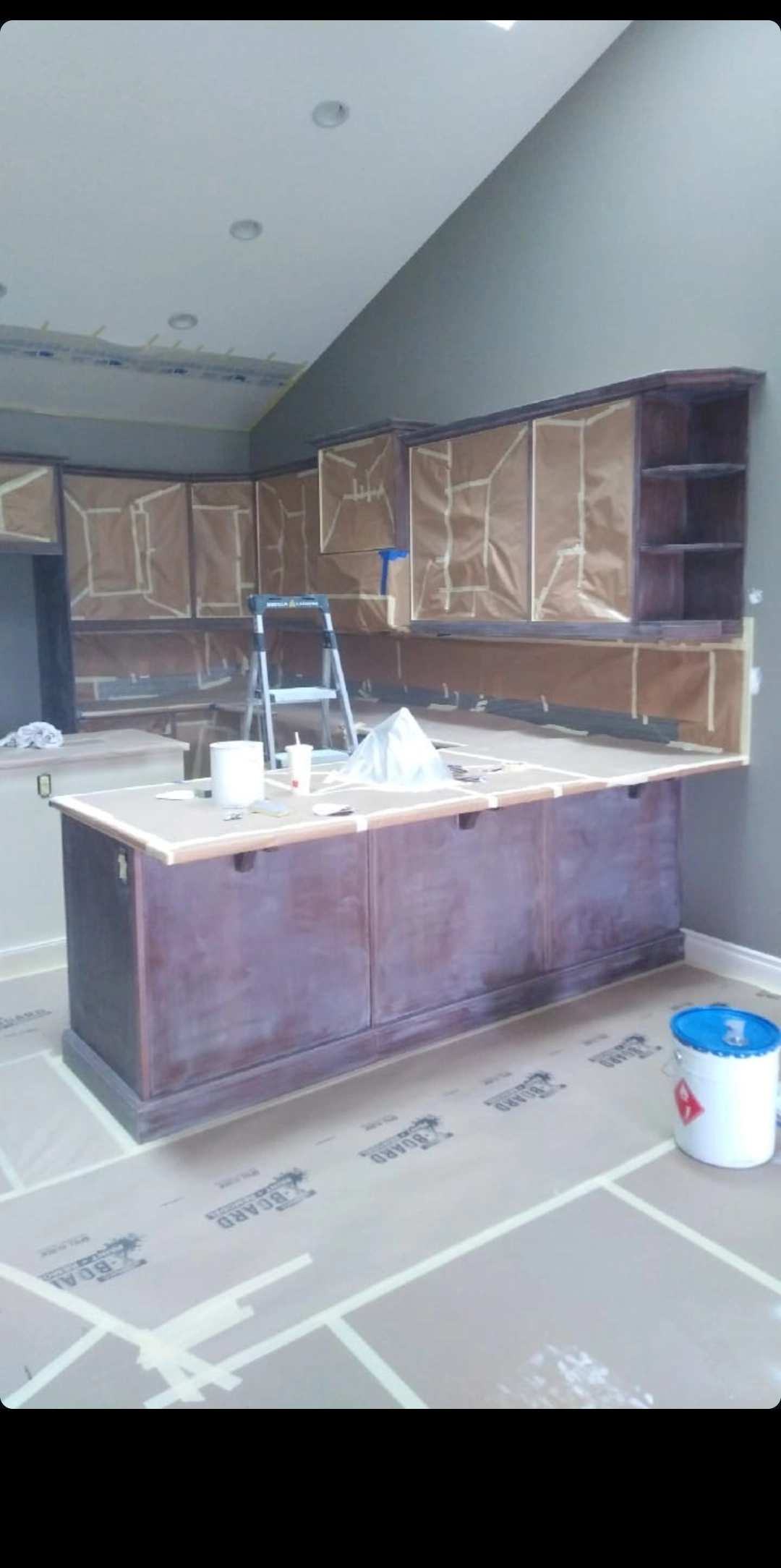 Golden Line kitchen cabinet project during preparation, with a wood peninsula, open corner shelves, paper-masked upper doors, a ladder and protected countertops and flooring